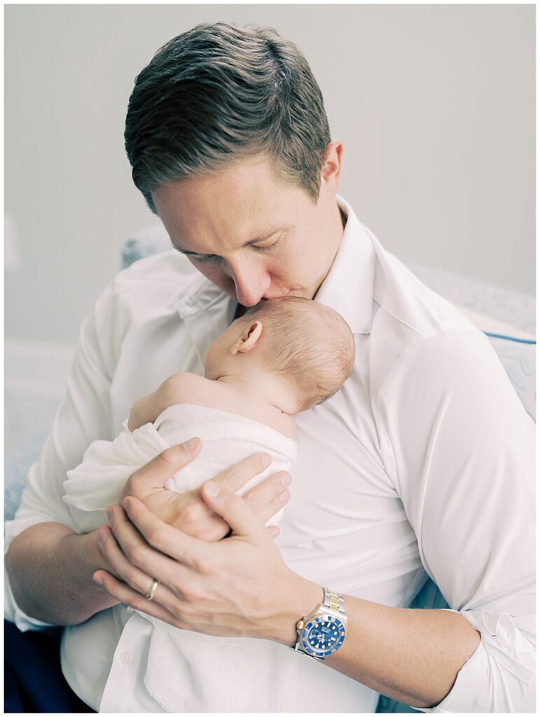 A blonde father leans down to kiss his baby's head while baby sleeps on his chest.