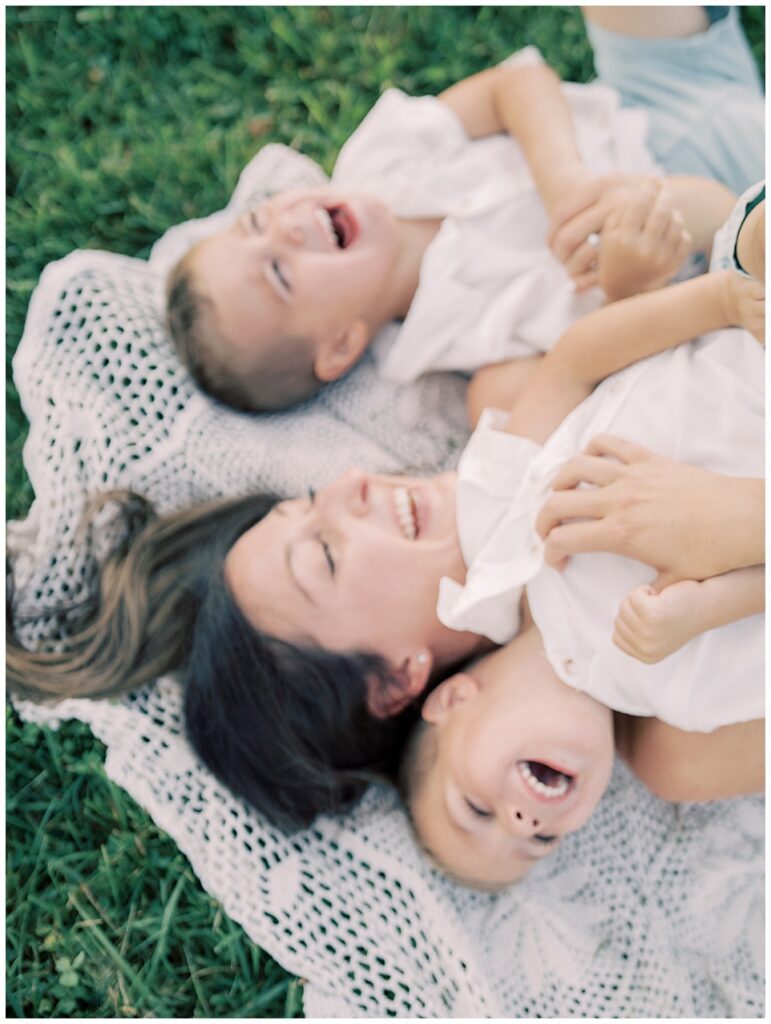 Out-of-focus image of mother with brown hair laughing with arms around her two twin boys while laying on crotchet blanket during Manassas Battlefield family session.