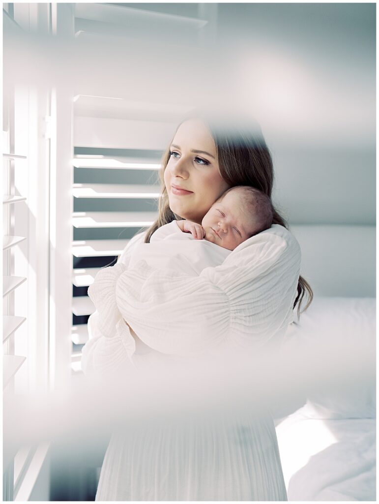 View of a mother with brown hair in a white dress holding her newborn baby through the white blinds in their bedroom during her North Bethesda newborn session.