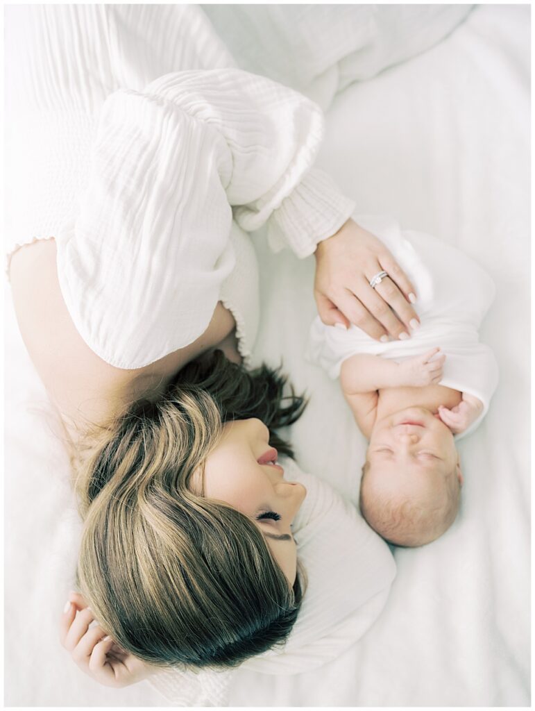 Mother with long brown hair lays on white bed with one hand on her newborn baby during her North Bethesda newborn session.