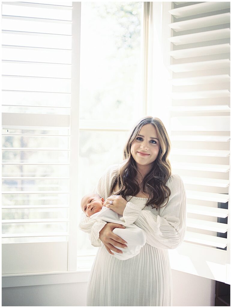 Long brown-haired mother holds her newborn baby girl while standing in front of blinds holding baby girl during her North Bethesda newborn session.