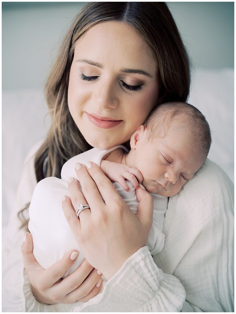 Brown hair mother holds newborn baby swaddled in white on her shoulder and closes her eyes during her North Bethesda newborn session.