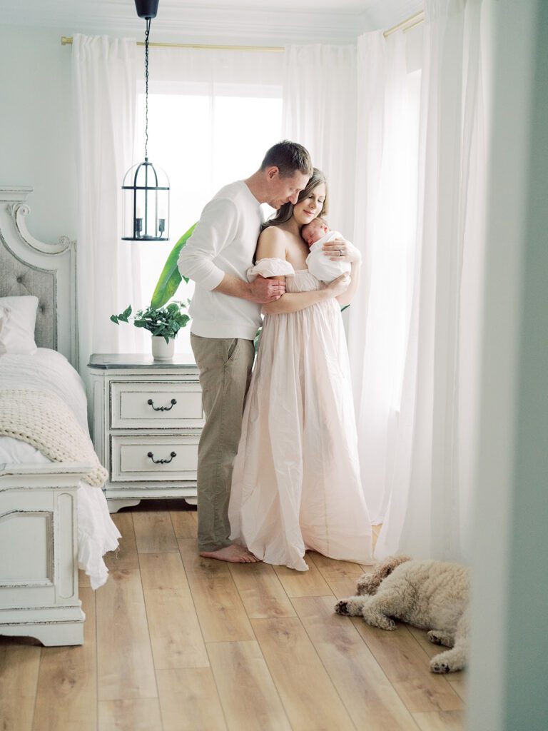 Mother and father stand cuddled together holding baby with dog laying nearby during their newborn photos in Annapolis.