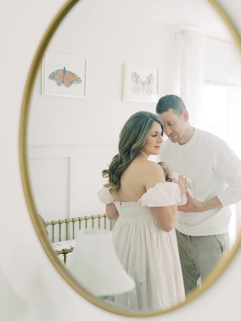 View from mirror of husband leaning into wife as they hold their newborn baby.