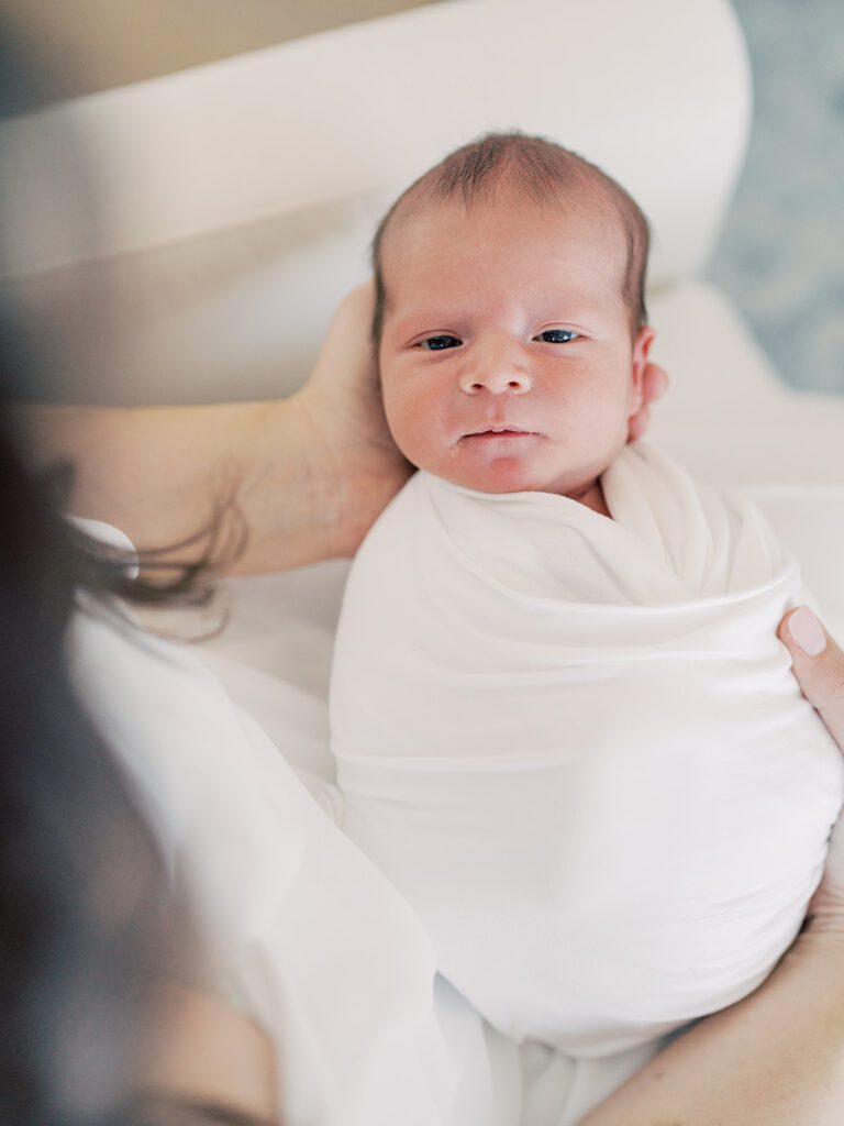 Baby swaddled in white gives a soft smile at the camera while being held by mother.