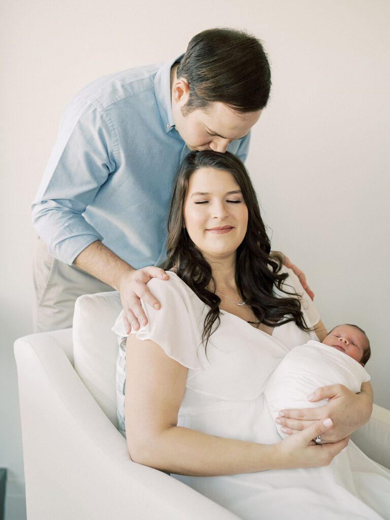 Father leans down to kiss the top of his wife's head as she sits in a chair and holds their newborn baby during their Georgetown newborn session.