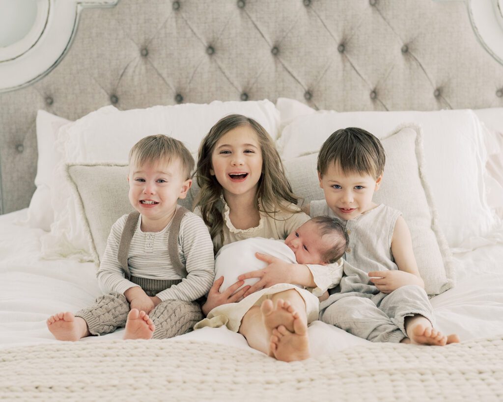 Big sister and two big brothers sit on bed holding newborn baby.