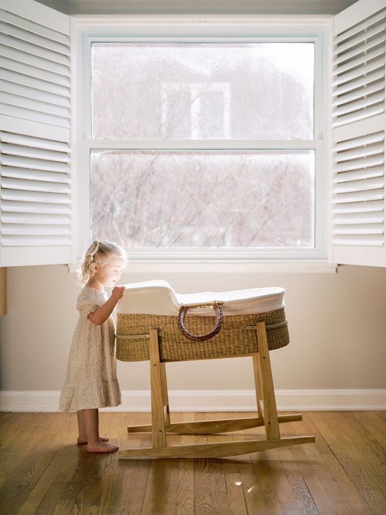 A little girl with blonde hair stands next to a bassinet in front of a window during their newborn photos in Arlington.