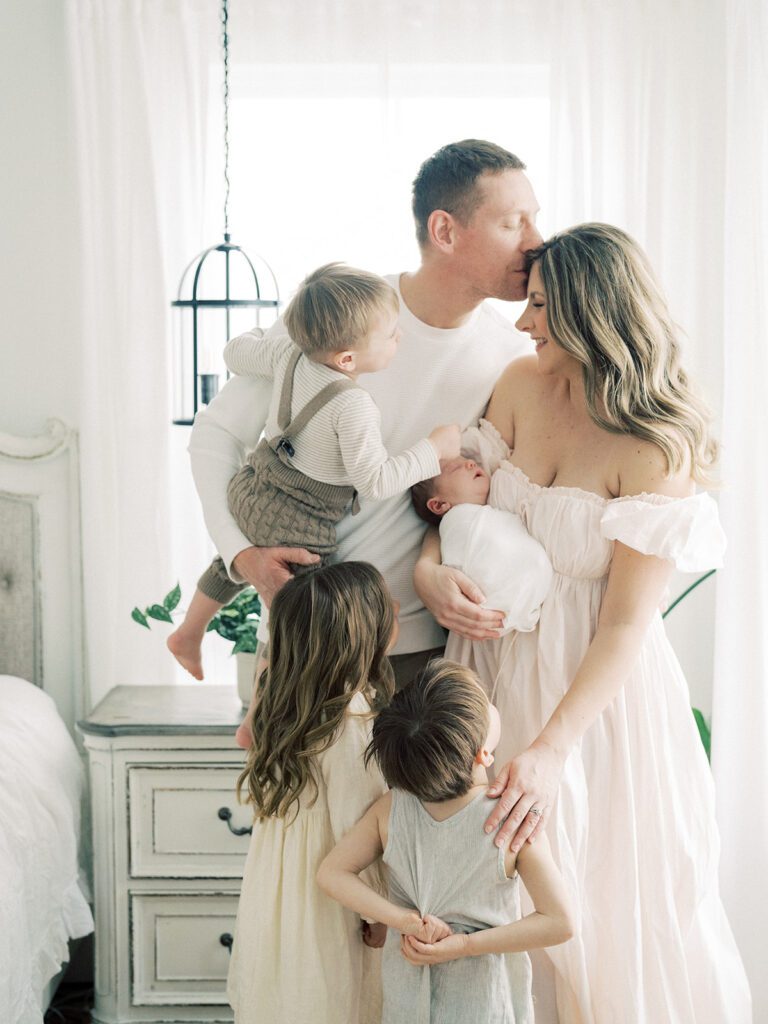 Father kisses wife on forehead as he holds young son and she holds newborn baby while their two other children stand close during their newborn photos in Annapolis.