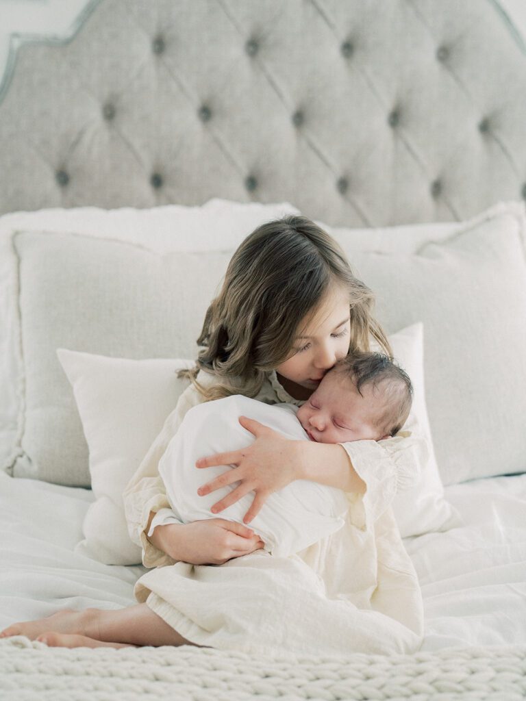 Big sister holds newborn baby while sitting on bed and kisses baby on head during their newborn photos in Annapolis.