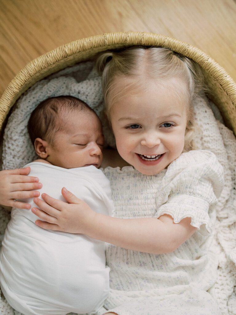 A blonde toddler holds her newborn brother in a bassinet during their newborn photos in Arlington.