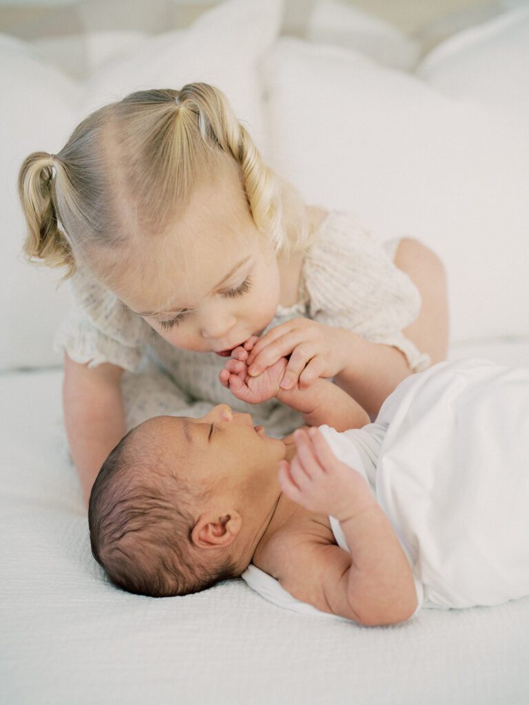 Blonde toddler girl in pigtails leans down to kiss her baby brother's fingers during their newborn photos in Arlington.