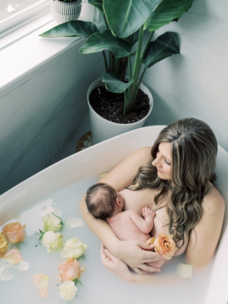 Overhead view of a mother sitting in a milk bath with her newborn daughter, surrounded by flowers.