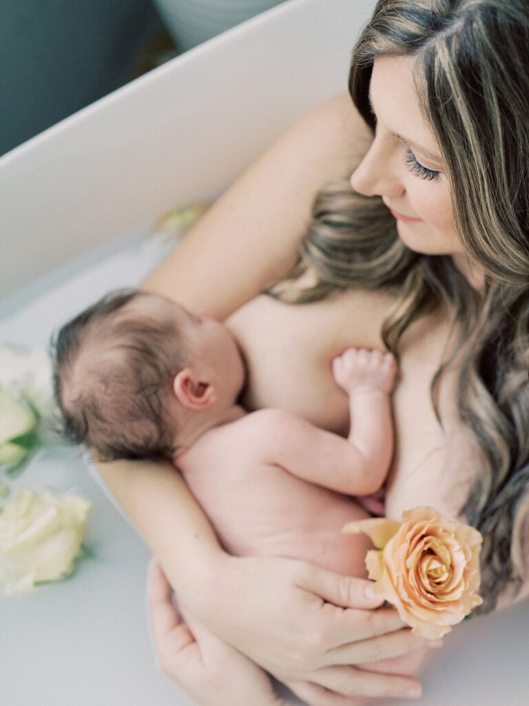 Mother with long brown hair looks down at her newborn daughter while holding her in a tub full of milk and flowers.