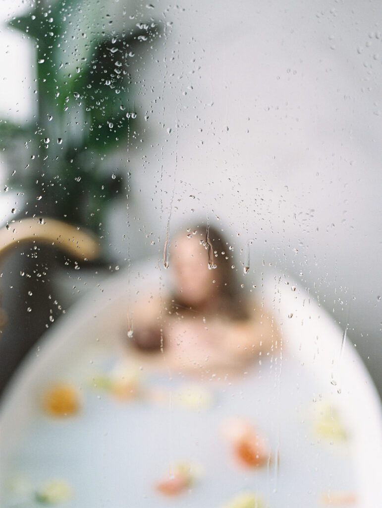 Milk bath photoshoots view of a glass door in a bathroom while a mother sits in a bath with her newborn daughter in the background.