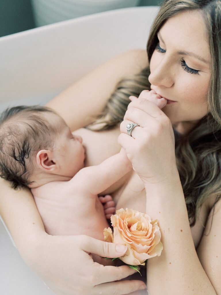 Mother kisses her daughter's fingers while holding her in a milk bath during their newborn session.