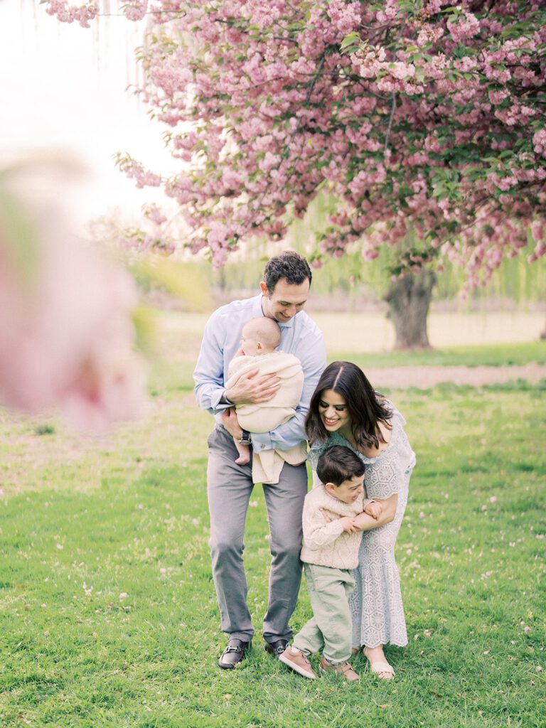 A mother leans down to hug her young son as she stands with her husband holding her baby daughter during their Kwanzan cherry blossom session.