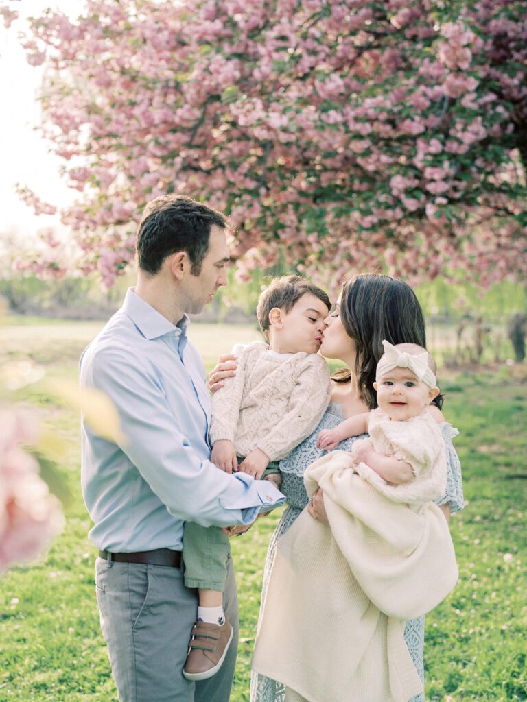 A little boy held by his father leans into his mother for a kiss during their family photo session with the Kwanzan cherry trees in DC.