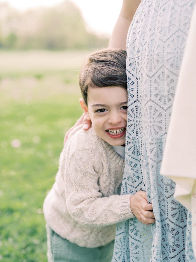 A little boy peeks behind his mother's legs and smiles.